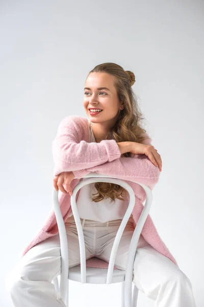 Chica sonriente sentada en una silla de madera blanca y mirando hacia otro lado aislada en blanco - foto de stock