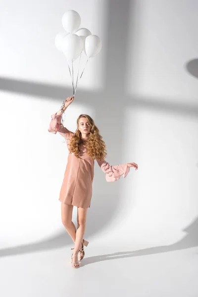 Beautiful woman in beige dress standing with hand up and holding bundle of balloons — Stock Photo