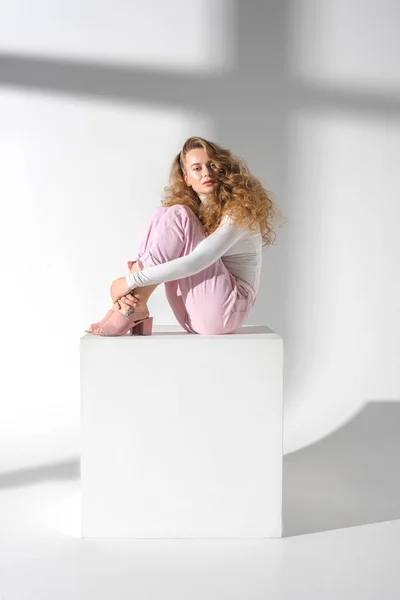 Belle femme sérieuse avec les cheveux bouclés assis sur le cube blanc et étreignant les jambes — Photo de stock