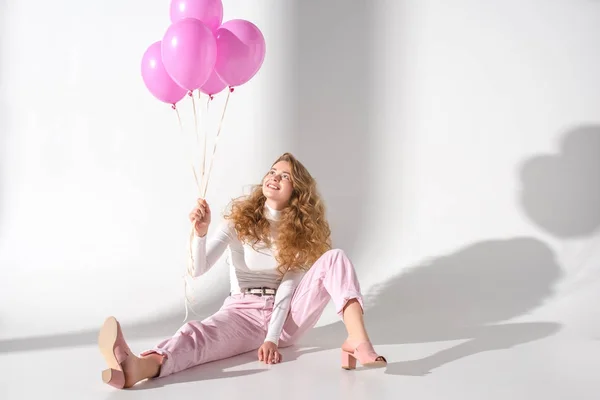 Sonriente chica con paquete de globos de color rosa sentado y mirando hacia arriba - foto de stock