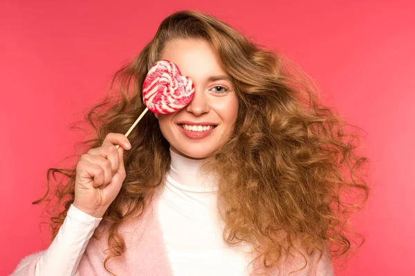 Smiling girl covering eye with heart shaped lollipop isolated on red — Stock Photo