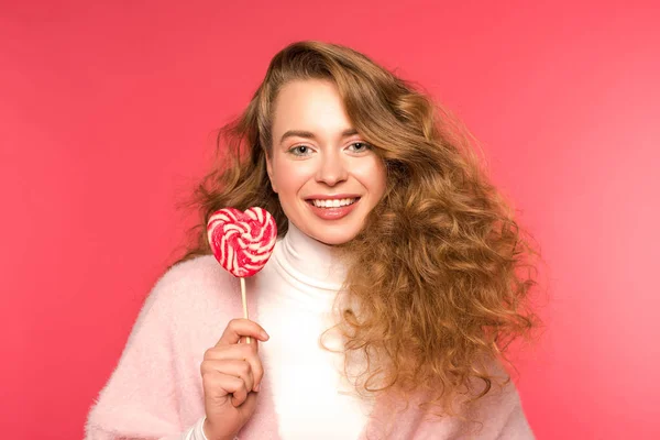 Mujer feliz de pie con piruleta en forma de corazón y mirando a la cámara aislada en rojo - foto de stock