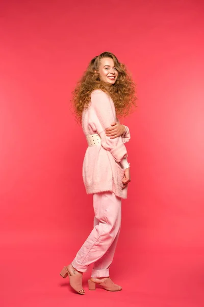 Стильная улыбающаяся девушка, стоящая в розовой одежде и смотрящая на камеру, изолированную на красный — стоковое фото