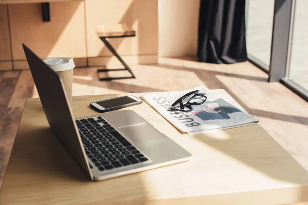 Laptop, smartphone, óculos e jornal de negócios na mesa no café — Fotografia de Stock