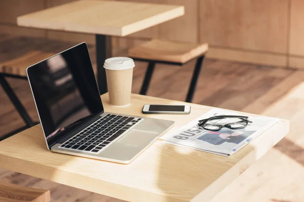 Laptop with blank screen, smartphone and business newspaper on table in coffee shop — Stock Photo