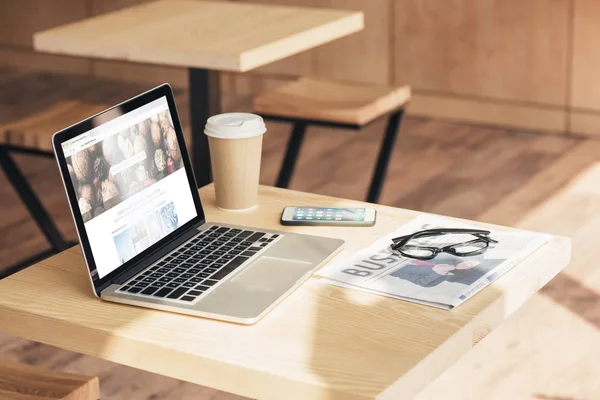 Laptop with depositphotos, smartphone and business newspaper on table in coffee shop — Stock Photo