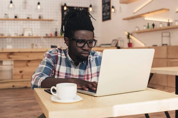 Hombre afroamericano usando el ordenador portátil en la cafetería con taza de café - foto de stock