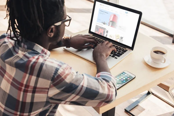 Hombre afroamericano usando el ordenador portátil con el sitio web de ebay y el teléfono inteligente - foto de stock