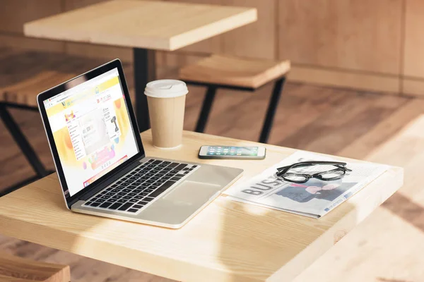 Laptop with aliexpress website, smartphone and business newspaper on table in coffee shop — Stock Photo