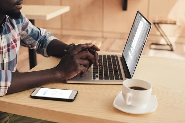 Vista recortada del hombre afroamericano utilizando teléfono inteligente con Facebook messenger y portátil con sitio web linkedin en la cafetería - foto de stock