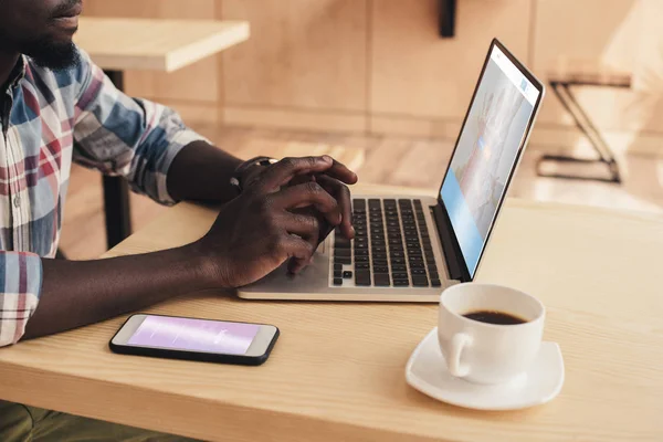 Vista cortada do homem americano africano usando smartphone com instagram e laptop com site de couchsurfing no café — Fotografia de Stock