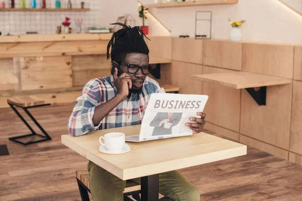 Homem americano africano falando no smartphone enquanto lê jornal de negócios no café — Fotografia de Stock
