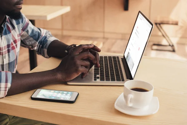 Vista recortada del hombre afroamericano utilizando el ordenador portátil con airbnb sitio web y teléfono inteligente en la cafetería - foto de stock