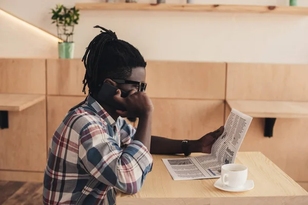 Homem americano africano falando no smartphone enquanto lê jornal no café — Fotografia de Stock