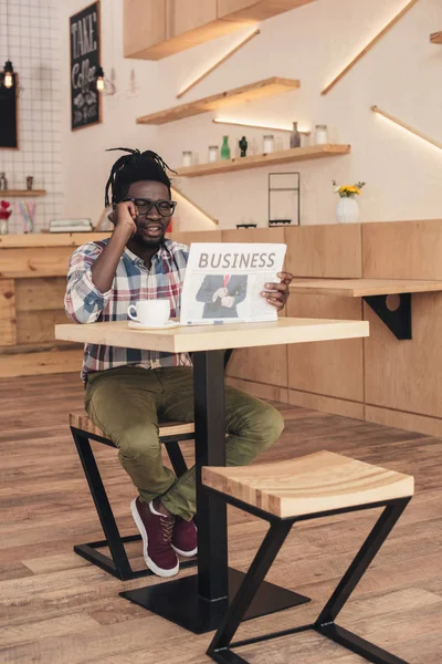 Африканский американец разговаривает на смартфоне, читая деловую газету в кафе — стоковое фото