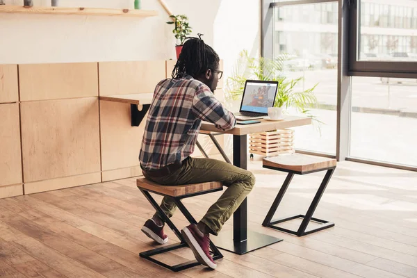 Vista posterior del hombre afroamericano utilizando el ordenador portátil con couchsurfing sitio web en la cafetería - foto de stock
