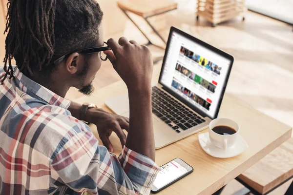 Hombre afroamericano usando el ordenador portátil con el sitio web de youtube y teléfono inteligente - foto de stock