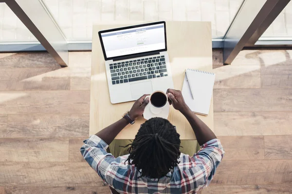 Vista aérea del hombre afroamericano bebiendo café y usando el ordenador portátil con el sitio web de facebook - foto de stock