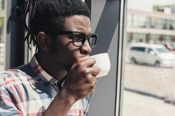 Africano americano uomo bere caffè mentre in piedi alla finestra — Foto stock