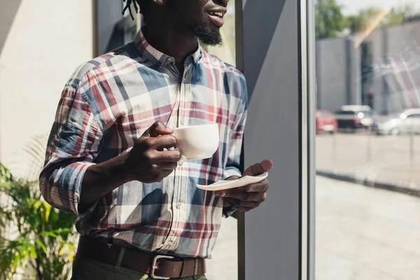 Vista ritagliata di uomo africano americano bere caffè mentre in piedi alla finestra — Foto stock