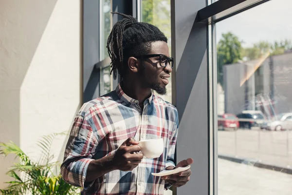 Africano americano hombre beber café mientras de pie en ventana - foto de stock