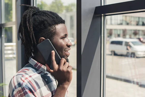 Cheerful african american man talking on smartphone while standing at window — Stock Photo