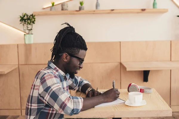 Homme afro-américain écrit dans le bloc-notes pendant la pause café dans le café — Photo de stock