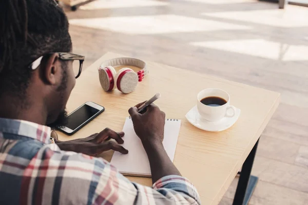 Homme afro-américain écrit dans le bloc-notes pendant la pause café dans le café — Photo de stock