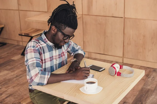 Lächelnder afrikanisch-amerikanischer Mann schreibt in Notizblock während Kaffeepause im Café — Stockfoto