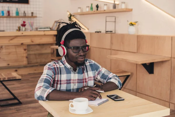 Pensativo afroamericano hombre escuchar música y escribir en bloc de notas en la cafetería - foto de stock