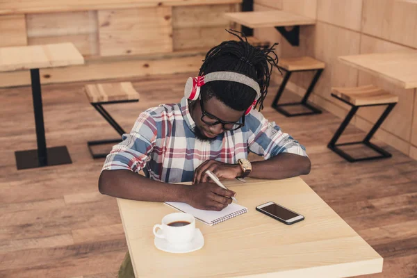Африканский американец слушает музыку и пишет в блокноте в кафе — стоковое фото