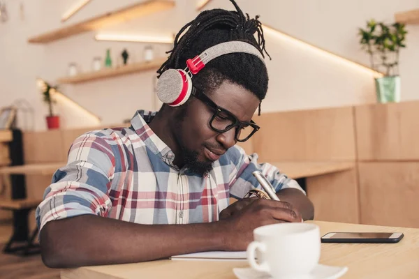 African american man listening music and writing in notepad in coffee shop — Stock Photo