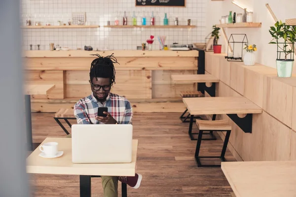 Hombre afroamericano usando el ordenador portátil y teléfono inteligente en la cafetería - foto de stock