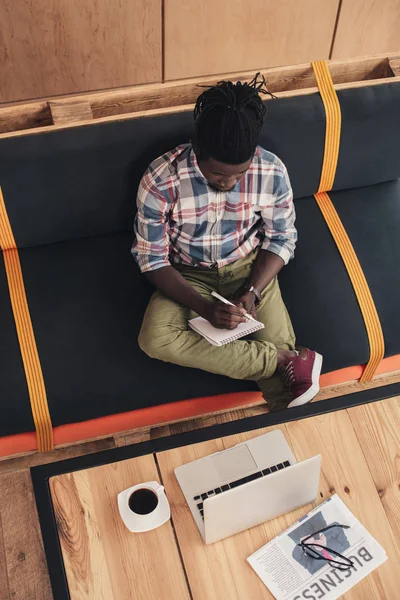Vue aérienne d'un homme afro-américain écrivant dans un bloc-notes assis dans un café avec un ordinateur portable et un journal d'affaires — Photo de stock