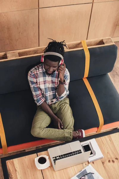 Vista aérea del hombre afroamericano escuchando música con auriculares y usando gadgets en la cafetería - foto de stock