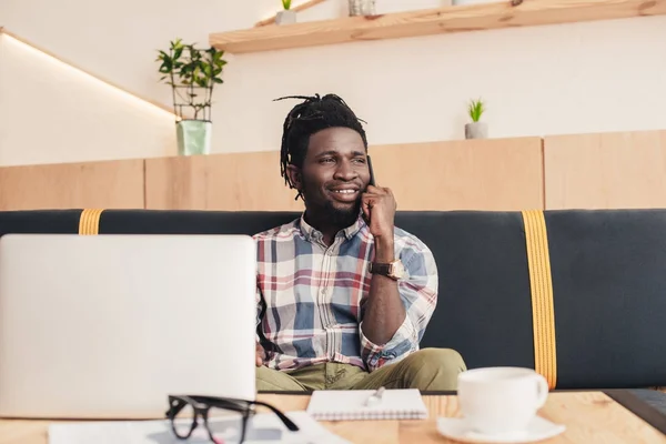 Sonriente hombre afroamericano usando teléfono inteligente y portátil en la cafetería - foto de stock