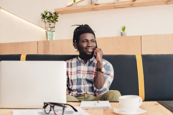 Uomo afroamericano sorridente utilizzando smartphone e laptop in caffetteria — Foto stock