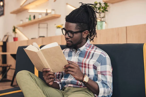 Hombre afroamericano leyendo libro mientras está sentado en el sofá en la cafetería - foto de stock
