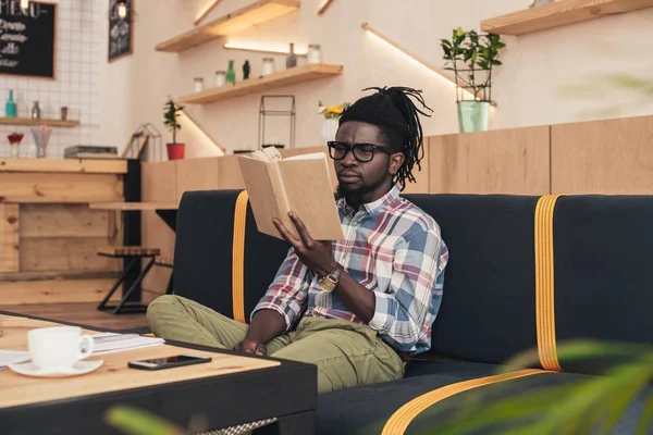 Афроамериканський чоловік читає книгу, сидячи на дивані в кафе — стокове фото