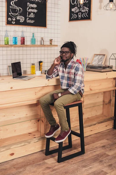 Hombre afroamericano usando el ordenador portátil y hablando en el teléfono inteligente en el mostrador de bar en la cafetería - foto de stock