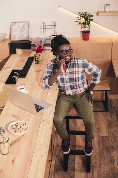 Веселый африканский американец, пользующийся ноутбуком и разговаривающий на смартфоне у барной стойки — стоковое фото