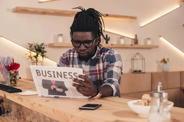 African american man reading business newspaper while sitting at bar counter with smartphone — Stock Photo