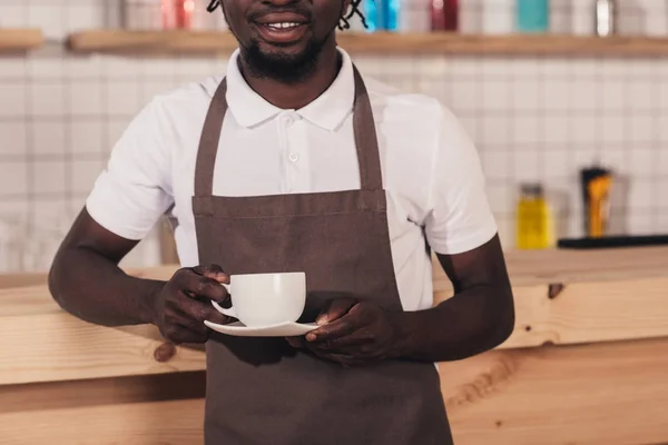 Vista cortada de barista americano africano em avental segurando xícara de café enquanto em pé no balcão bar — Fotografia de Stock