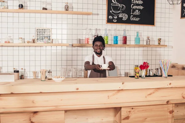 African american barista in apron holding coffee cup while standing at bar counter — Stock Photo