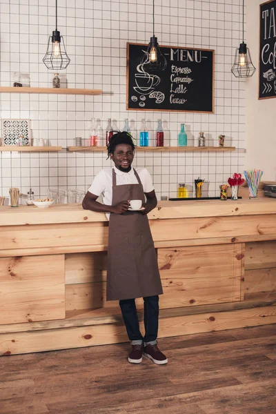 Африканский американский бариста в фартуке держа чашку кофе, стоя у барной стойки — стоковое фото