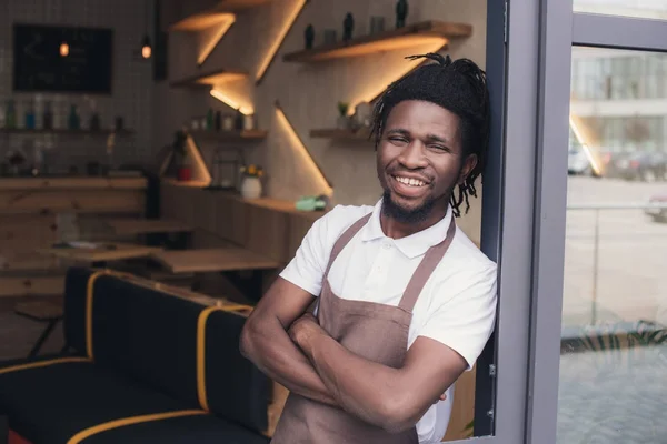 Cheerful african american cafe owner in apron standing at entrance — Stock Photo