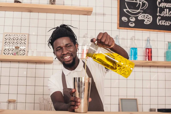 Sonriente barman afroamericano haciendo cóctel en coctelera en el mostrador del bar - foto de stock