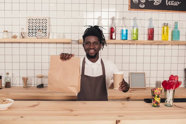 Barista afroamericano sosteniendo taza de café desechable y paquete kraft en el mostrador de bar - foto de stock