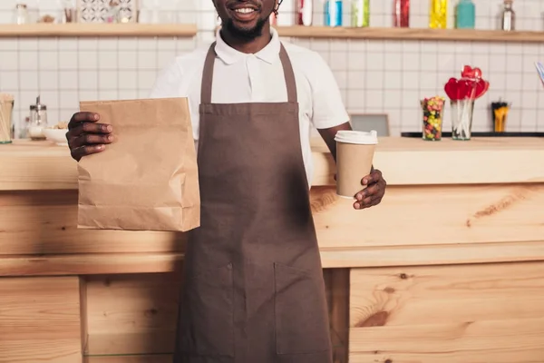 Cropped view of african american barista holding disposable cup of coffee and kraft package at bar counter — Stock Photo