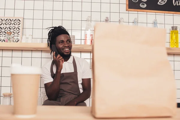 African american barista talking on smartphone, disposable cup of coffee and kraft package standing on foreground on bar counter — Stock Photo